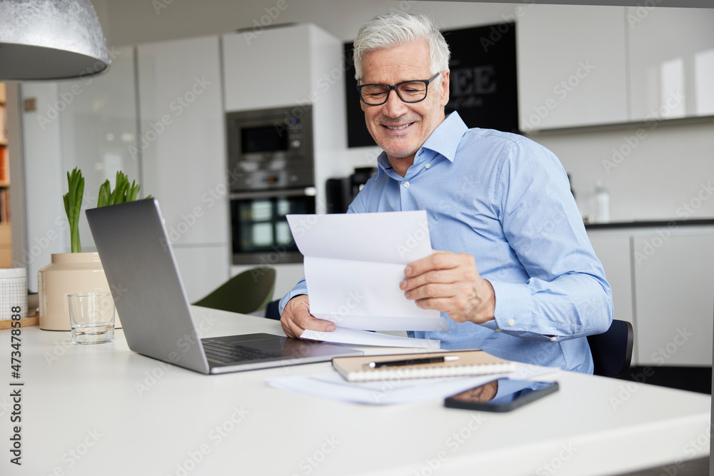Smiling male entrepreneur reading document while sitting with laptop in office Smiling male entrepreneur reading document while sitting with laptop in office