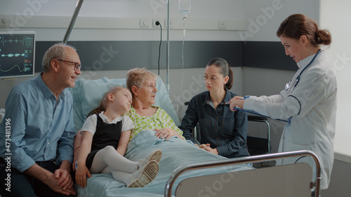 Sick grandma and family talking to physician about healthcare in hospital ward. Medical specialist explaining disease to senior patient in bed while she has visitors at clinic. Checkup visit photo