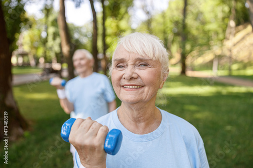 Cheerful senior lady does exercises with dumbbell training with partner in park