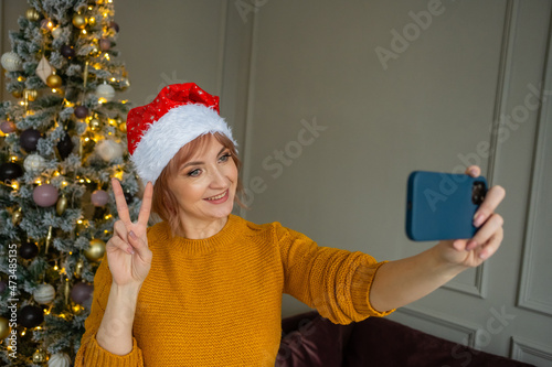 Happy woman in santa claus hat takes a selfie on their smart phone at christmas tree at home. Female are calling perents and friends through video call to congratulate them on new year. photo