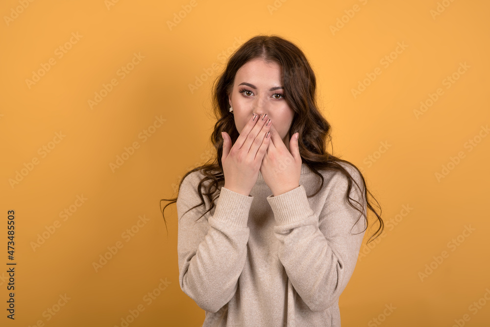 Surprised and shocked caucasian brunette girl with wavy hair in a casual jumper covering her mouth with her hands on an orange studio background. Surprised and shocked caucasian brunette girl with wavy hair in a casual jumper covering her mouth with her hands on an orange studio background.