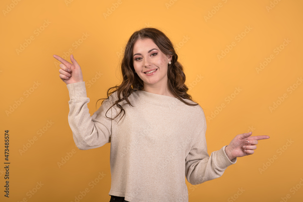 Happy and attractive Caucasian brunette girl with wavy hair in a casual jumper showing index fingers to the sides on an orange studio background. Happy and attractive Caucasian brunette girl with wavy hair in a casual jumper showing index fingers to the sides on an orange studio background.