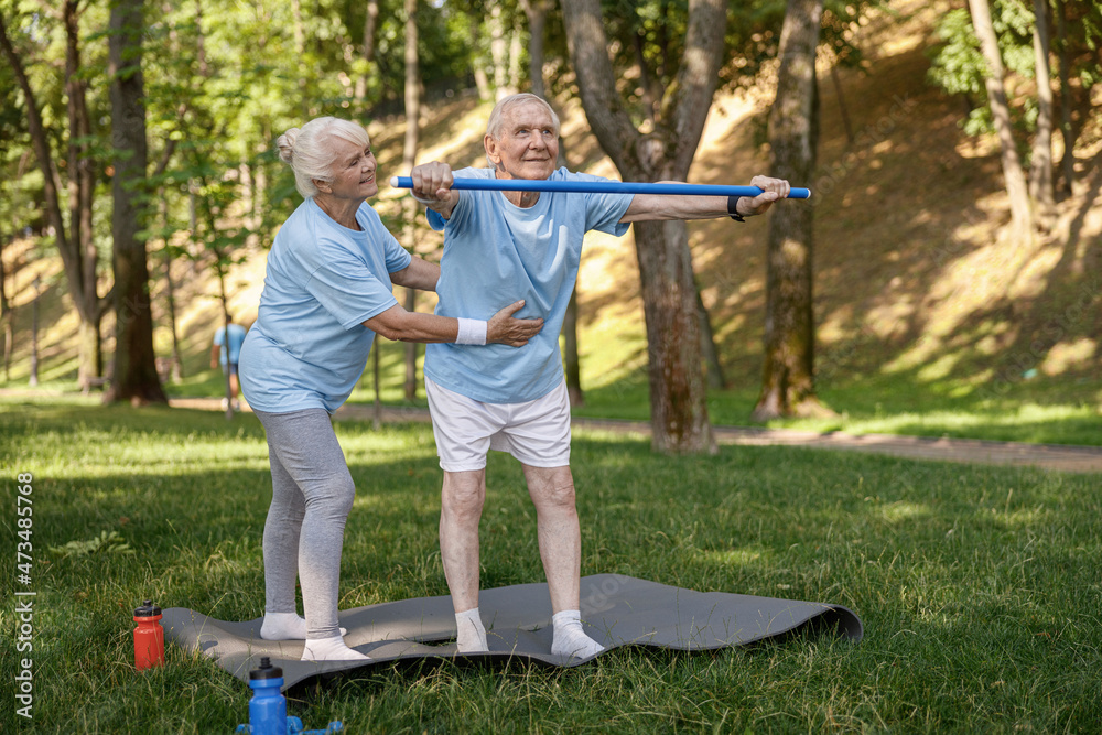 Smiling woman helps senior patient to do exercise with bar in picturesque park Smiling woman helps senior patient to do exercise with bar in picturesque park
