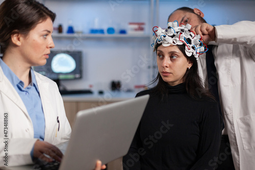 Neurologist doctor holding laptop computer showing tomography to woman patient while researcher man adjusting eeg headset during neuroscience experiment. Team of researchers analyzing brain evolution photo