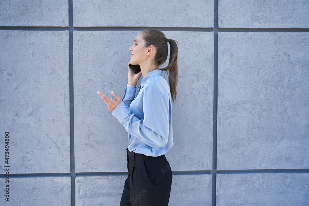 Businesswoman talking on mobile phone while walking by wall Businesswoman talking on mobile phone while walking by wall