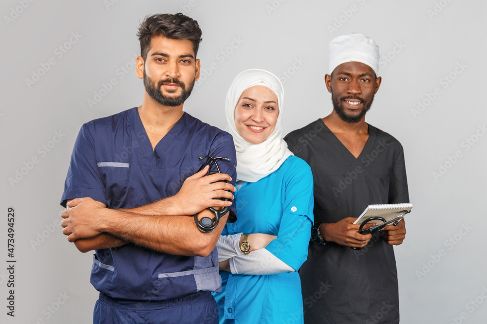 Portrait of positive healthcare workers standing in the hallway and looking at the camera. Group of medics smiling over gray background. muslim doctor woman on background of african and indian doctor Portrait of positive healthcare workers standing in the hallway and looking at the camera. Group of medics smiling over gray background. muslim doctor woman on background of african and indian doctor