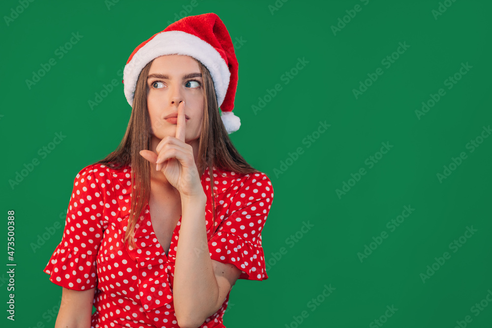 Portrait of a cheerful woman in Santa outfit with a finger on her lips. A young girl shows a gesture of silence. Secret concept of Christmas or New Year Portrait of a cheerful woman in Santa outfit with a finger on her lips. A young girl shows a gesture of silence. Secret concept of Christmas or New Year