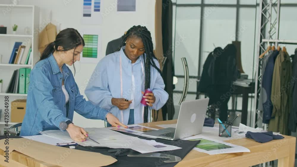 Fashion Designers, a Caucasian Woman and an African-American Woman, Create a New Fashion Collection By Working With Sketches, Fabrics, Using a Laptop in the Studio. Apparel Design Startup Concept. Fashion Designers, a Caucasian Woman and an African-American Woman, Create a New Fashion Collection By Working With Sketches, Fabrics, Using a Laptop in the Studio. Apparel Design Startup Concept.