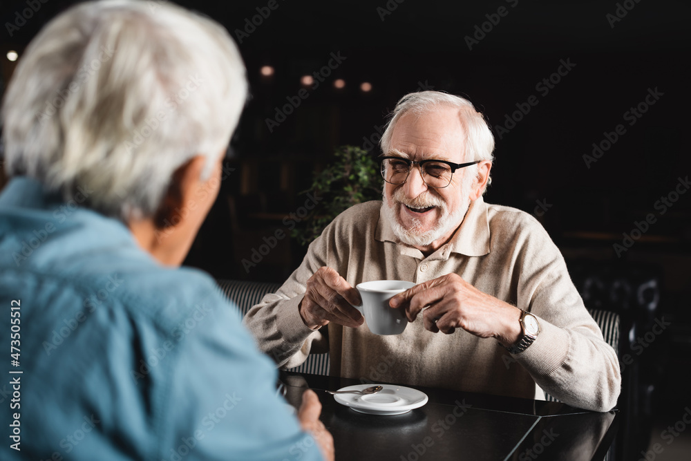 joyful senior man in eyeglasses holding coffee cup near blurred friend in bar joyful senior man in eyeglasses holding coffee cup near blurred friend in bar