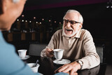 cheerful senior man in eyeglasses holding cup of coffee while touching hand of blurred friend in cafe