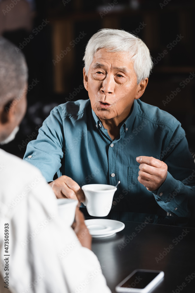 senior asian man holding cup of coffee while talking to blurred african american friend senior asian man holding cup of coffee while talking to blurred african american friend