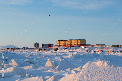The helicopter flies over the northern coastal settlement. Winter arctic landscape. Ice hummocks and snow. Buildings and satellite dishes on the shore. Tavayvaam, Chukotka, the Far North of Russia.