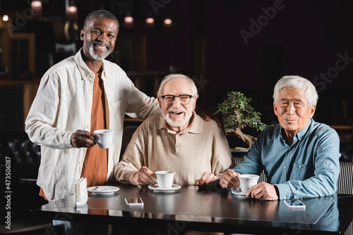 elderly multiethnic friends smiling at camera while drinking coffee in restaurant
