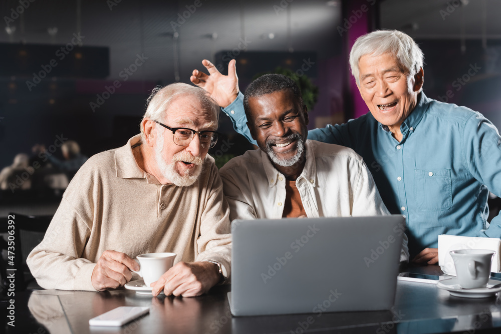 cheerful asian man waving hand near senior multiethnic friends during video call on laptop in cafe cheerful asian man waving hand near senior multiethnic friends during video call on laptop in cafe