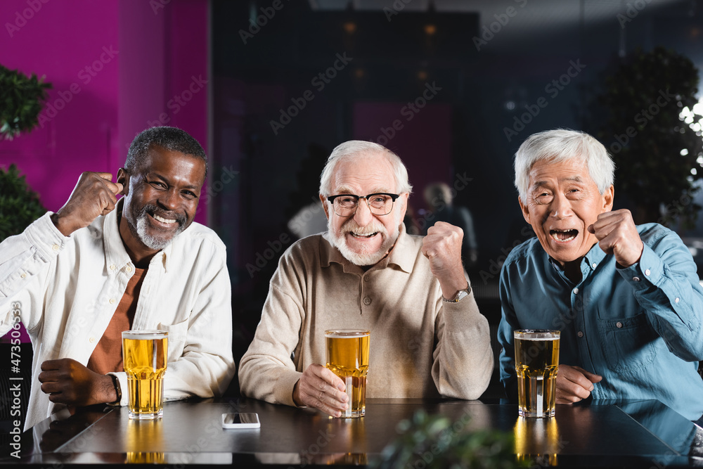 happy senior multiethnic friends showing win gesture while watching football championship in pub