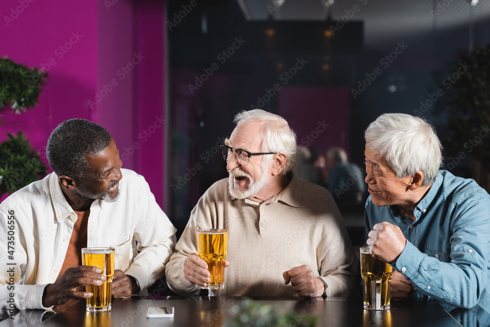 excited elderly multicultural friends talking in pub near glasses of beer