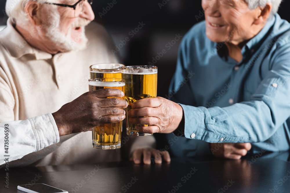 senior multiethnic friends clinking glasses of beer while spending time in pub on blurred background