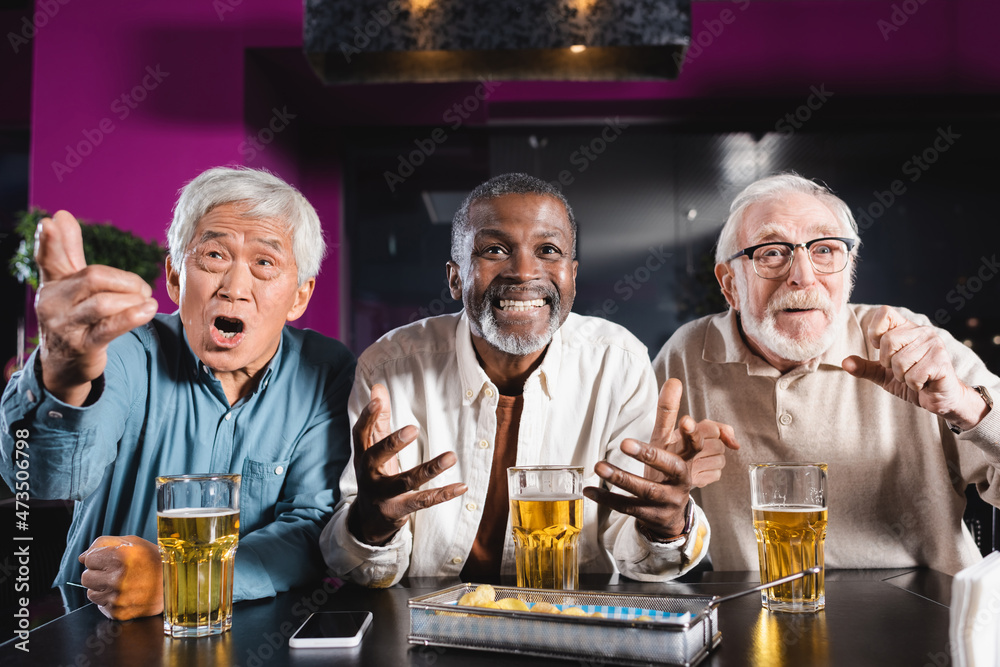 emotional senior multicultural friends watching football match near glasses of beer and chips in pub