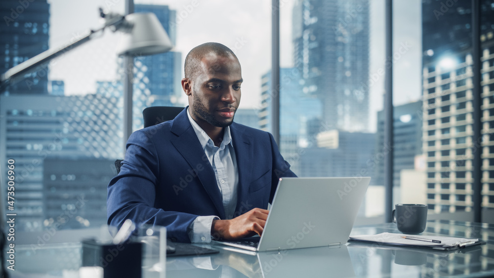 Fototapeta premium Successful Black Businessman in Tailored Suit Working on Laptop Computer on Top Floor Office Overlooking Big City. Professional CEO Managing Environmental, Social and Corporate Governance