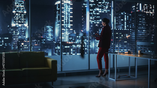 Successful Powerful Businesswoman Wearing Stylish Suit Holding Laptop Standing in Big City Night Office. Female CEO Managing Environmental, Social and Corporate Governance for e-Commerce Empire photo