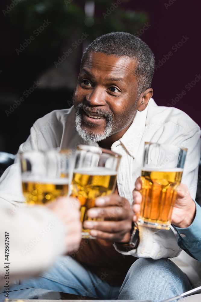 senior african american man smiling while clinking blurred beer glasses with friends senior african american man smiling while clinking blurred beer glasses with friends