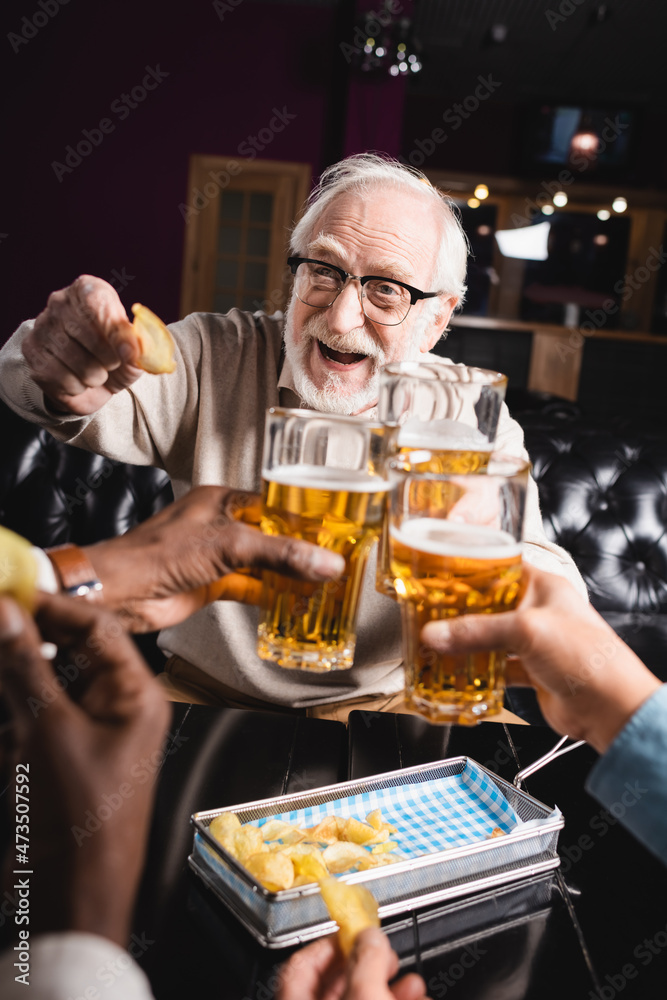 cheerful elderly man holding chips while clinking beer glasses with blurred friends