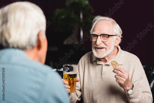 smiling elderly man holding glass of beer and chips near blurred friend in pub
