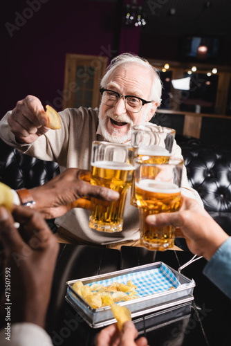 cheerful elderly man holding chips while clinking beer glasses with blurred friends