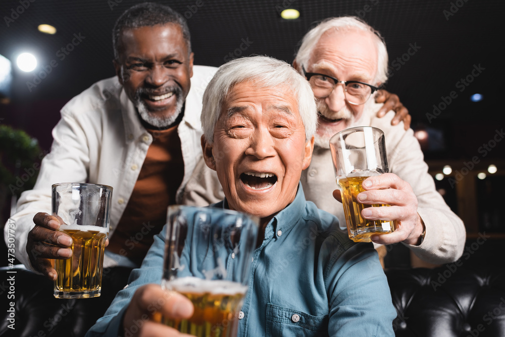 cheerful multiethnic friends holding glasses with beer while looking at camera in pub