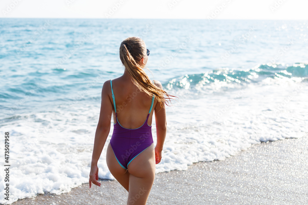 An attractive young woman comes out of the sea in a swimsuit. A girl in a swimsuit walks under the bright summer sun along the sand of the Mediterranean coast. Rest by the sea. Selective focus An attractive young woman comes out of the sea in a swimsuit. A girl in a swimsuit walks under the bright summer sun along the sand of the Mediterranean coast. Rest by the sea. Selective focus