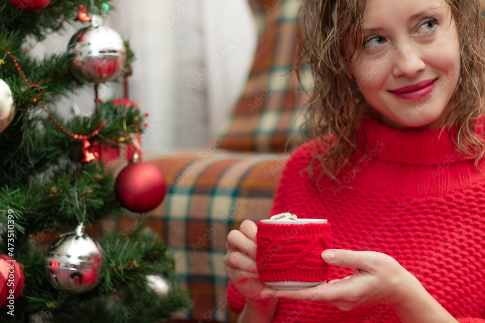 Young beautiful woman in a red sweater with a mug of cocoa and marshmallows at home by the new year tree on new year's eve. Selective focus. Portrait