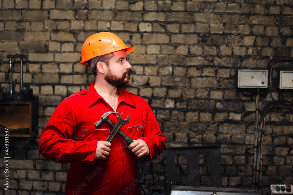 Profile portrait of handsome bearded technician or worker wearing red overalls and orange hard hat holding hammer and wrench at factory workshop