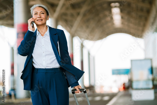 Grey asian woman talking on cellphone while walking with suitcase