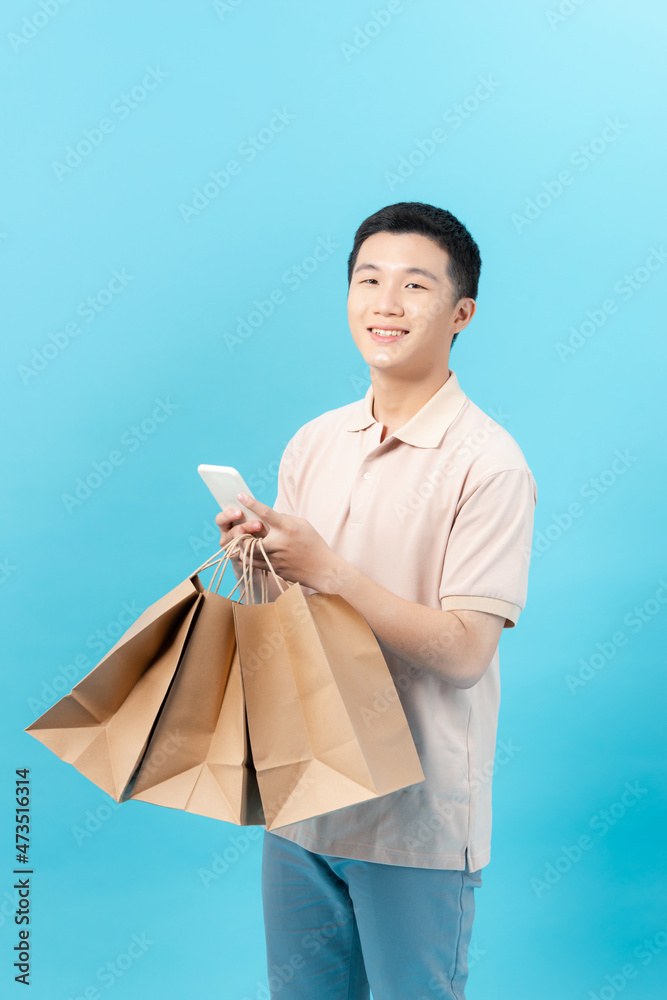 Young handsome southeast Asian man holding mobile phone and shopping bags in light blue studio isolated background