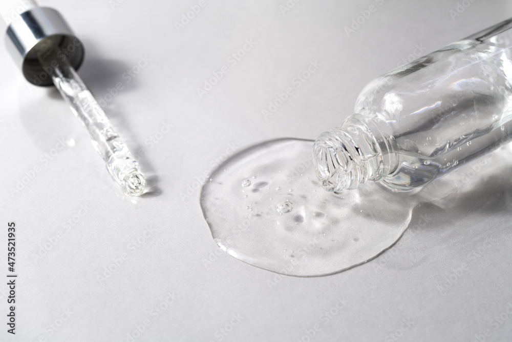 Transparent facial serum pouring from glass bottle and pipette on grey background. Liquid gel for skin care. Cosmetic product with hyaluronic acid closeup. Beauty routine.