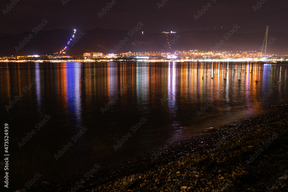 Night city with lights and reflections in the water and mountains near the sea and red sky, coast in pebbles, very beautiful at night in travel photography.