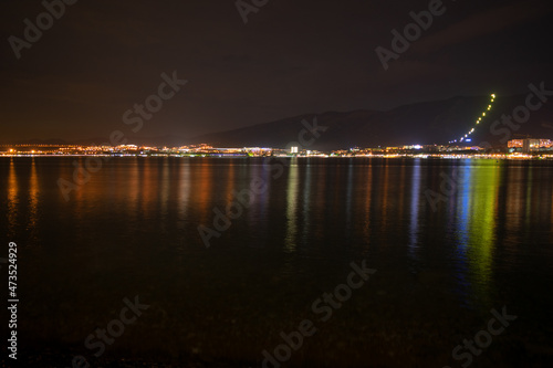 Night city with lights and reflections in the water and mountains by the sea and red sky, pebble shore, very beautiful at night on a journey.
