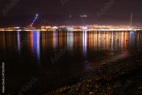 Fototapeta Naklejka Na Ścianę i Meble -  Night city with lights and reflections in the water and mountains near the sea and red sky, coast in pebbles, very beautiful at night in travel photography.