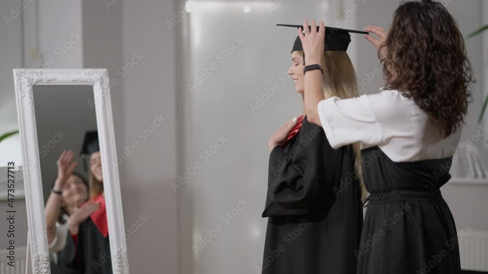 Side view slim young woman in graduation outfit admiring reflection in mirror as mother adjusting hair and clothes. Happy confident Caucasian graduate getting dressed for ceremony with parent helping