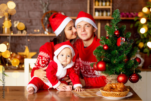 New Year or Christmas, close-up of a baby in a red sweater and Santa Claus hat in the kitchen, a happy young family mom, dad and baby at the Christmas tree smiling, hugging