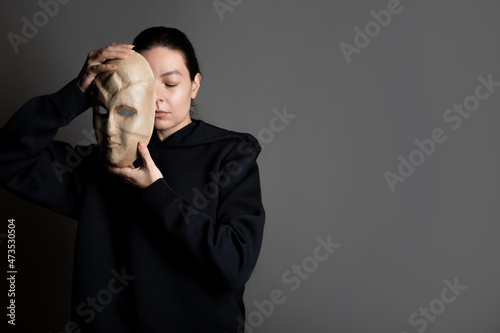 Obraz na plátně Hiding behind a mask, a young woman in a dark hoodie hides her face with a mask, self-identification problems and impostor syndrome