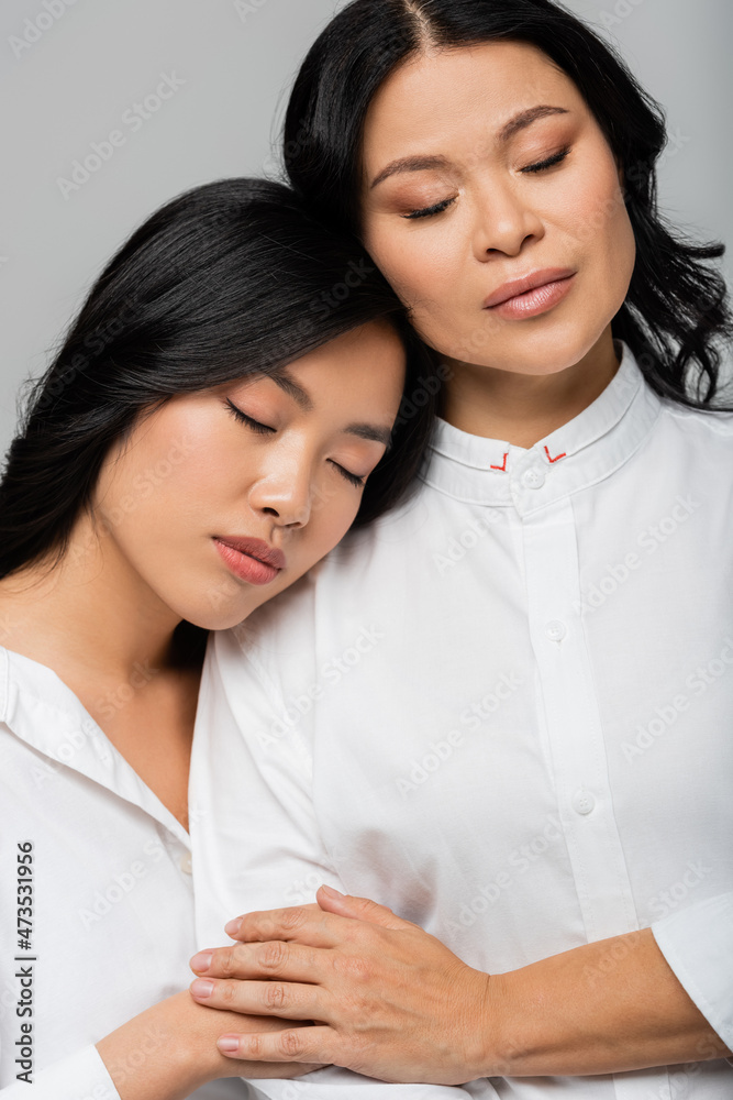 asian young adult daughter leaning on shoulder of mother with closed eyes isolated on grey asian young adult daughter leaning on shoulder of mother with closed eyes isolated on grey