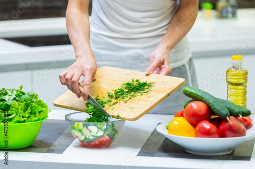 Close up of a girl hands cutting greenery with a knife on a cutting board for a vegan vitamin vegetable salad and putting them in a glass bowl while cooking breakfast in the kitchen