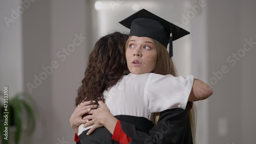 Young female graduate in gown and hat hugging woman with dissatisfied irritated facial expression. Caucasian hypocritical woman embracing mother on graduation day. Egoism concept photo