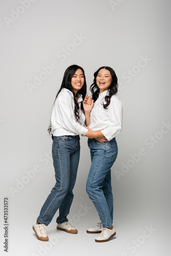 full length of cheerful asian mother and young adult daughter in denim jeans and white shirts posing on grey