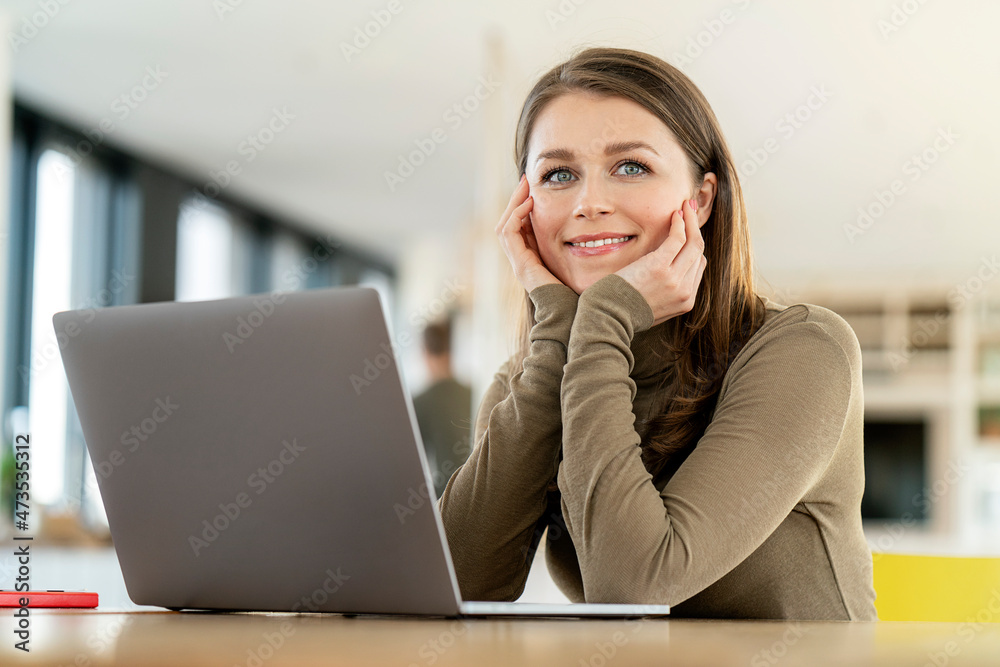 Smiling businesswoman with hand on chin looking away while sitting in front of laptop at office Smiling businesswoman with hand on chin looking away while sitting in front of laptop at office