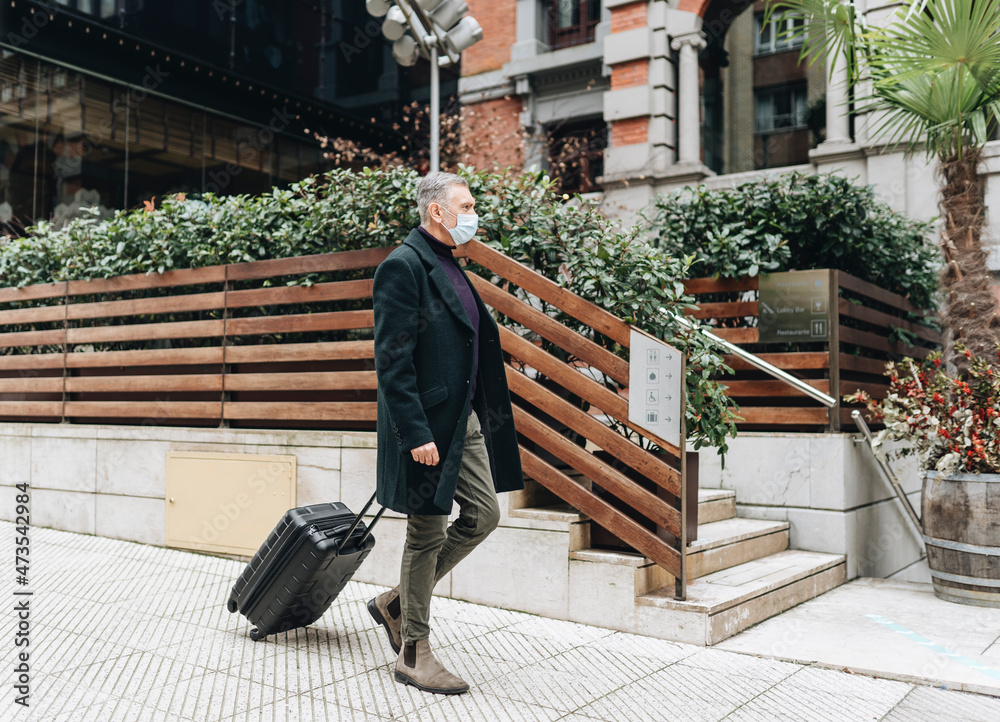 Mature man wearing protective face mask walking with luggage on footpath Mature man wearing protective face mask walking with luggage on footpath
