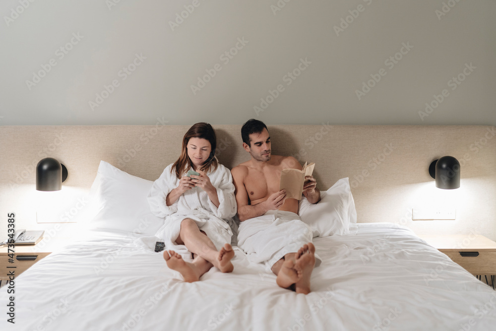 Mid adult couple reading book and using smart phone on bed Mid adult couple reading book and using smart phone on bed