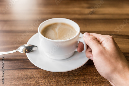 A man takes a cup of coffee on a saucer with his hand against the background of a wooden table in a cafe