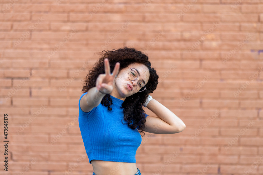 Woman with hand in hair showing peace gesture in front of wall Woman with hand in hair showing peace gesture in front of wall
